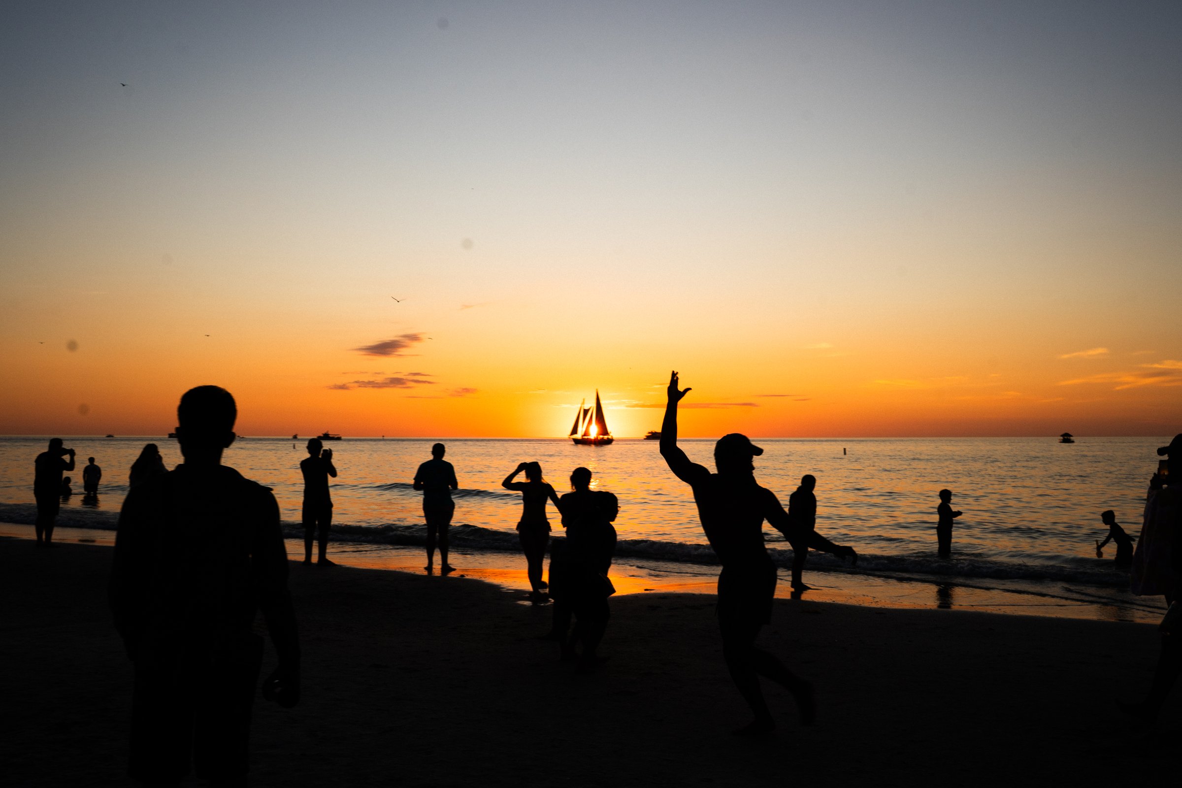 Silhouetted figures on beach at vibrant orange sunset with sailboat on the horizon